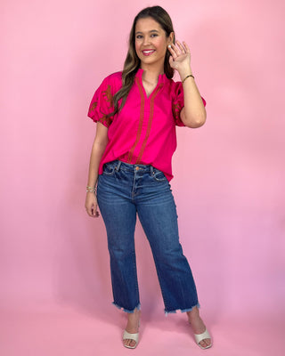 Woman wearing a pink blouse with red embroidery and blue jeans on a pink background