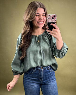 Woman taking a mirror selfie wearing a green blouse and blue jeans on a gold background