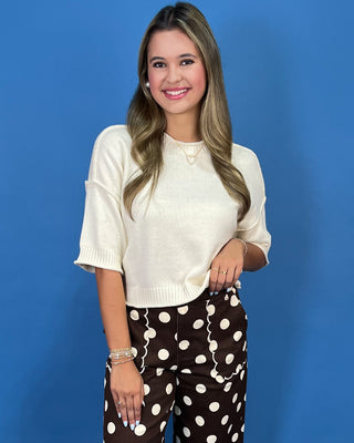 A woman posing against a solid blue background, wearing a cream-colored knit short-sleeve top and brown pants with white polka dots.