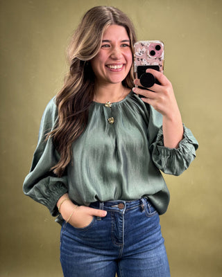 Woman taking a selfie wearing a green blouse and blue jeans against a gold background