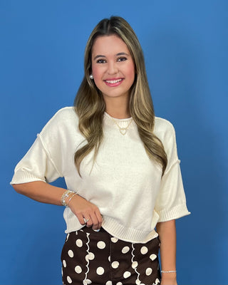 A woman with long blonde-highlighted hair smiling, wearing a cream short-sleeve knit top and brown polka-dot pants with white wavy lines, standing against a solid blue background.