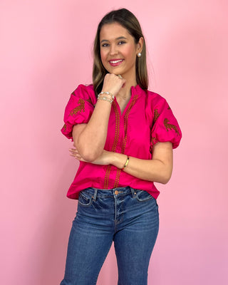 Woman wearing a pink embroidered blouse and blue jeans against a pink background