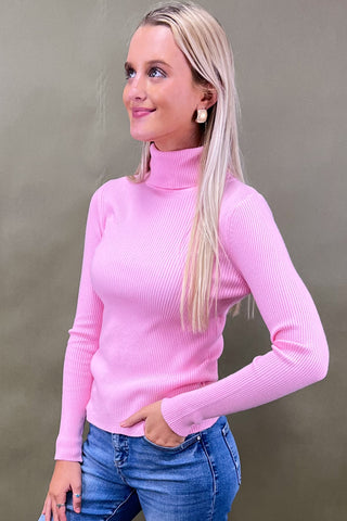 Woman wearing a pink turtleneck sweater and blue jeans against a plain background