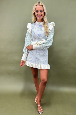 Woman wearing a pastel floral knit sweater dress with ruffle trim and puff sleeves, styled with strappy nude heels, boutique fashion photography on olive green backdrop