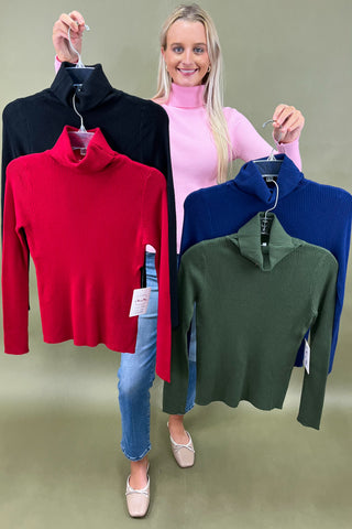 Woman holding four different colored turtleneck sweaters on hangers against a plain background.