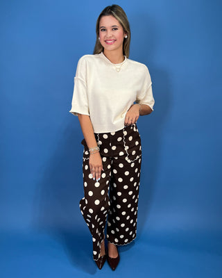 A woman modeling a cream short-sleeve knit top and brown wide-leg pants with white polka dots, standing against a solid blue studio backdrop.