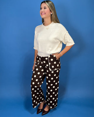 A woman modeling a cream short-sleeve knit top and brown wide-leg pants with white polka dots, standing against a solid blue studio background.
