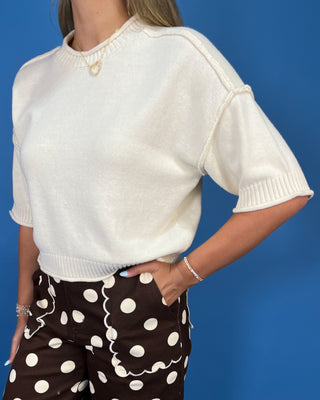 A woman modeling a cream short-sleeve knit top and brown wide-leg pants with white polka dots, standing against a solid blue studio background.