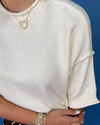 A close-up of a person wearing a cream-colored knit short-sleeve sweater with ribbed trim, accessorized with a heart-shaped necklace and bracelets, set against a solid blue background.
