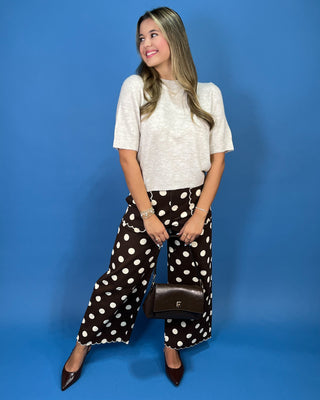 A woman posing in a white short-sleeve knit top and black wide-leg pants with white polka dots, holding a small brown leather handbag against a solid blue studio background.