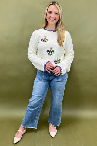 Woman wearing a white sweater with colorful patterns and blue jeans against a green background