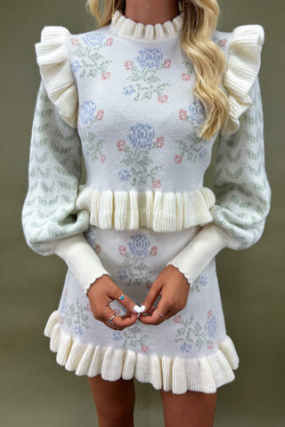 Woman wearing a pastel floral knit sweater dress with ruffle trim and puff sleeves, styled with strappy nude heels, boutique fashion photography on olive green backdrop