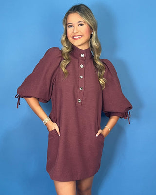 Burgundy mini shift dress with a high neckline, statement jeweled buttons, puff sleeves with tie cuffs, and convenient side pockets.