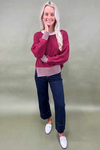 Woman wearing a red sweater, striped shirt, blue jeans, and white shoes on a plain background