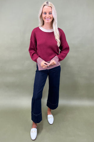 Woman wearing a burgundy sweater and dark blue jeans against a plain background