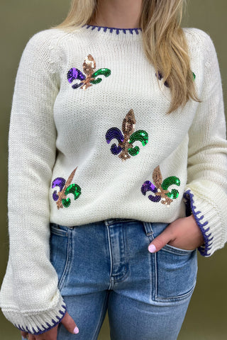 Person wearing a white sweater with colorful designs and blue jeans against a neutral background