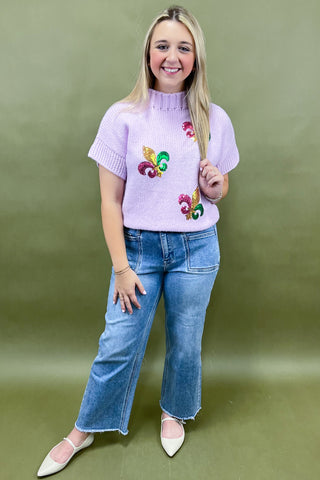 Woman wearing a light purple sweater with floral designs and blue jeans against a green background