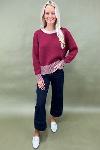 Woman wearing a burgundy sweater and dark blue jeans against a plain background