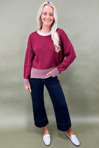 Woman wearing a burgundy sweater, navy pants, and white shoes on a plain background