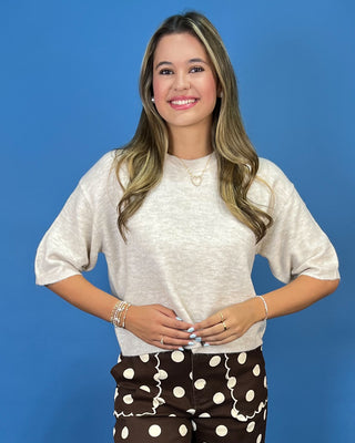 A woman modeling a light beige short-sleeve knit top and brown polka-dot pants against a solid blue studio background.