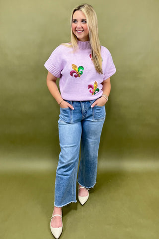 Woman wearing a light purple sweater with colorful designs and blue jeans against a green background