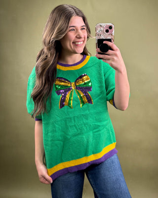 Woman taking a selfie wearing a green sweater with a colorful bow design on a beige background