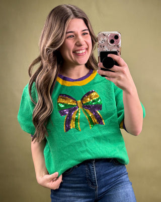 Woman wearing a green sweater with a colorful bow design, taking a selfie against a beige background.