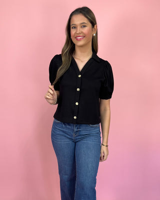 Woman wearing a black blouse with short sleeves and blue jeans against a pink background