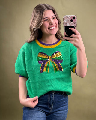 Woman wearing a green sweater with a colorful bow design, taking a selfie against a beige background.