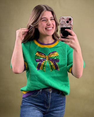 Person wearing a green sweater with a colorful bow design, taking a selfie against a plain background.