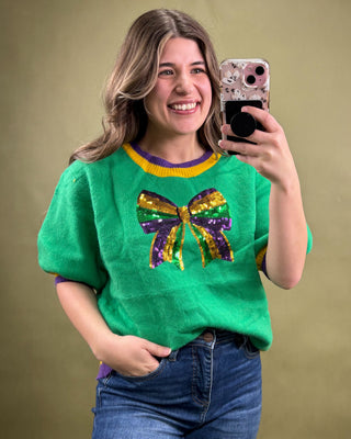 Woman wearing a green sweater with a colorful bow design, taking a selfie against a beige background.