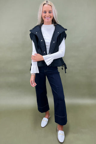 Black sherpa zip vest layered over white ribbed long sleeve top styled with dark wide leg cropped jeans and white loafers.