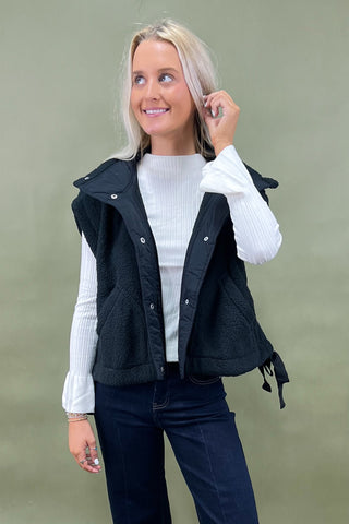 Black sherpa zip vest layered over white ribbed long sleeve top styled with dark wide leg cropped jeans and white loafers.