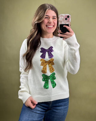 Woman wearing a white sweater with colorful sequin bows, taking a selfie against a beige background.
