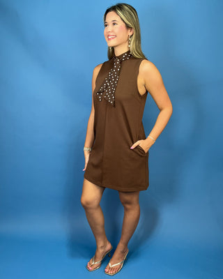 A woman modeling a sleeveless brown mini dress with a high neck and a matching brown polka-dot neck scarf