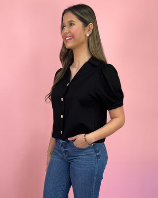 Woman wearing a black blouse with puff sleeves and blue jeans against a pink background
