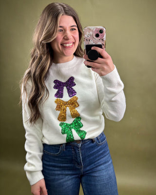 Person wearing a white sweater with colorful bow designs, taking a selfie against a plain background.