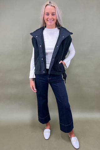 Black sherpa zip vest layered over white ribbed long sleeve top styled with dark wide leg cropped jeans and white loafers.
