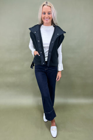 Black sherpa zip vest layered over white ribbed long sleeve top styled with dark wide leg cropped jeans and white loafers.
