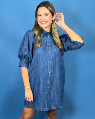 A woman modeling a blue denim shirt dress with puff sleeves, a button-down front, and side pocket