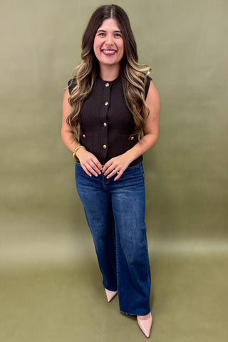 Woman wearing a black sleeveless top with gold buttons and blue jeans on a green background