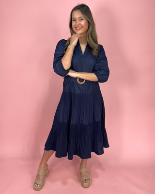 Woman wearing a navy blue dress with pleated layers on a pink background