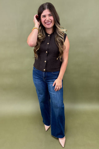 Woman wearing a black sleeveless top with gold buttons and blue jeans on a green background