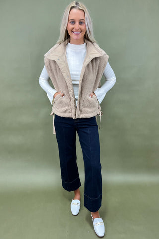 Woman wearing a beige vest over a white shirt, dark jeans, and white shoes on a green background