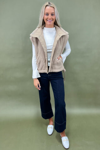 Woman wearing a beige vest over a white shirt with dark jeans on a green background