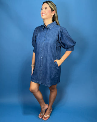 A woman modeling a blue denim puff-sleeve shirt dress against a solid blue studio backdrop.