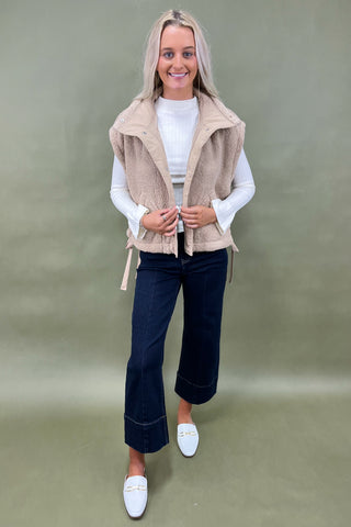 Woman wearing a beige vest, white shirt, dark jeans, and white shoes on a green background