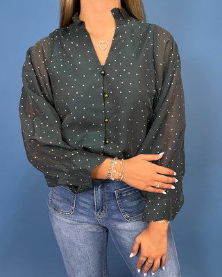A woman modeling a dark green sheer long-sleeve blouse with small white polka dots, paired with blue jeans and metallic flats, standing against a solid blue studio backdrop.