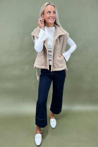 Woman wearing a beige vest over a white long-sleeve shirt, dark jeans, and white loafers on a green background