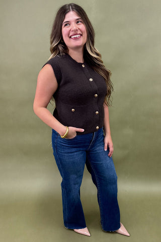 Woman wearing a black sleeveless top with gold buttons and blue jeans against a green background